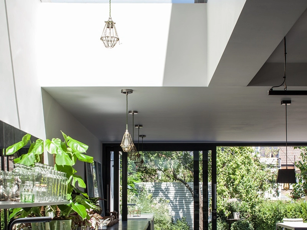 Modern kitchen featuring a sleek black countertop, stylish pendant lights, and large glass doors opening to a lush green garden. Natural light streams in through a skylight, enhancing the airy atmosphere.