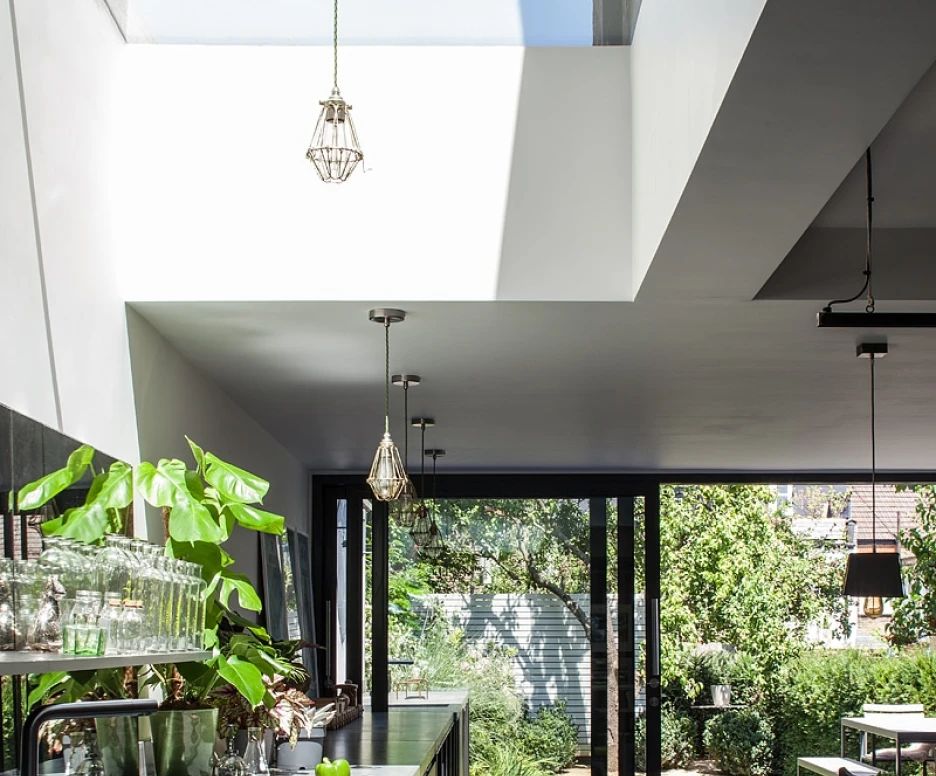 Modern kitchen featuring a sleek black countertop, stylish pendant lights, and large glass doors opening to a lush green garden. Natural light streams in through a skylight, enhancing the airy atmosphere.