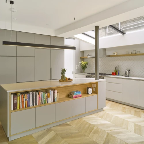 Cocina moderna con una isla central de madera, estanterías con libros y una encimera clara. Muebles en tonos grises y detalles en madera. Ventanales que permiten la entrada de luz natural.