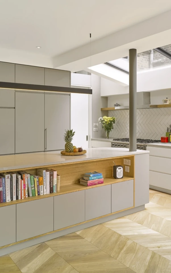 Moderne Küche mit hellen, minimalistischen Schränken und einer großen Kücheninsel. Auf der Insel stehen Bücher und eine Ananas. Der Boden ist mit hellen Holzdielen ausgelegt, und große Fenster sorgen für viel Licht.