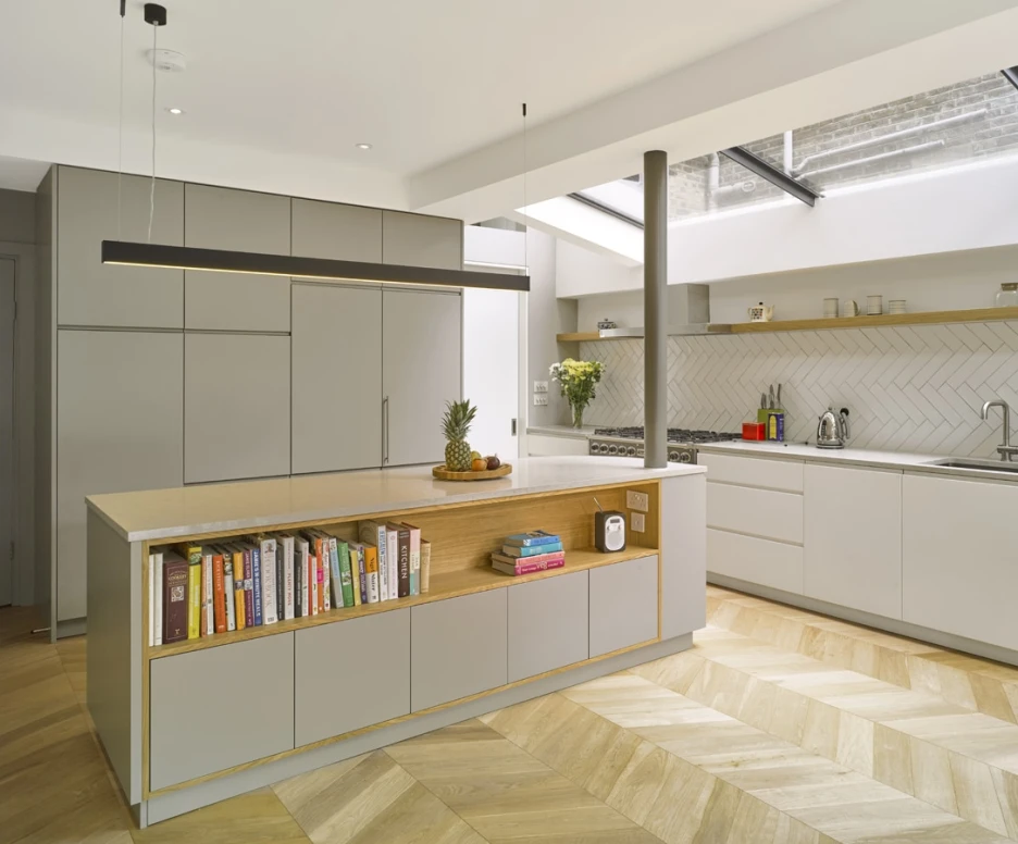 Cuisine moderne avec un îlot central en bois et un espace de rangement. Étagères ouvertes avec des livres de cuisine. Éclairage naturel provenant d’une fenêtre de toit. Sol en bois à motifs chevron.