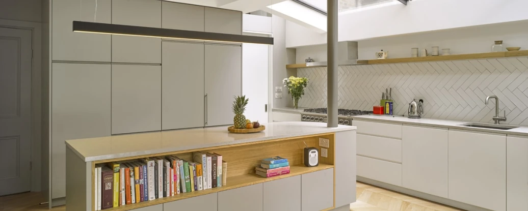 Modern kitchen featuring a large island with a wood-accented shelf displaying books, and sleek gray cabinetry. The space is illuminated by natural light from a skylight, with herringbone-patterned wood flooring and minimalist decor.