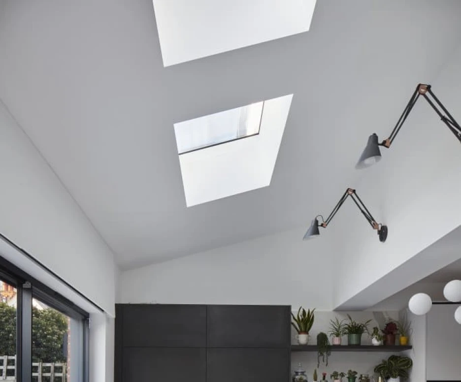 A modern kitchen featuring sleek white cabinets, a black island with a marble vein design, and skylights above. The space has minimalistic decor, with plants on shelves and stylish lighting fixtures. Large glass doors lead to an outdoor area.