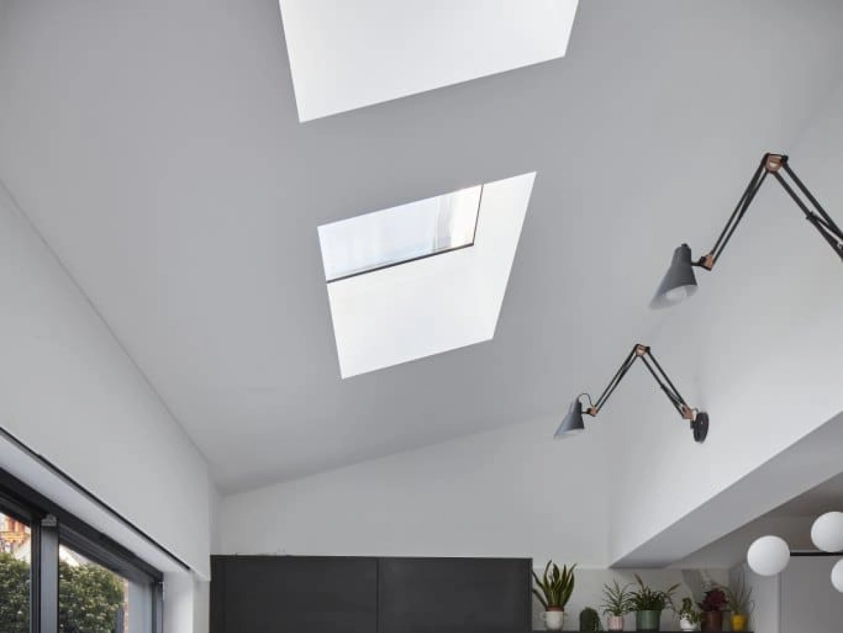 A modern kitchen featuring sleek white cabinets, a black island with a marble vein design, and skylights above. The space has minimalistic decor, with plants on shelves and stylish lighting fixtures. Large glass doors lead to an outdoor area.