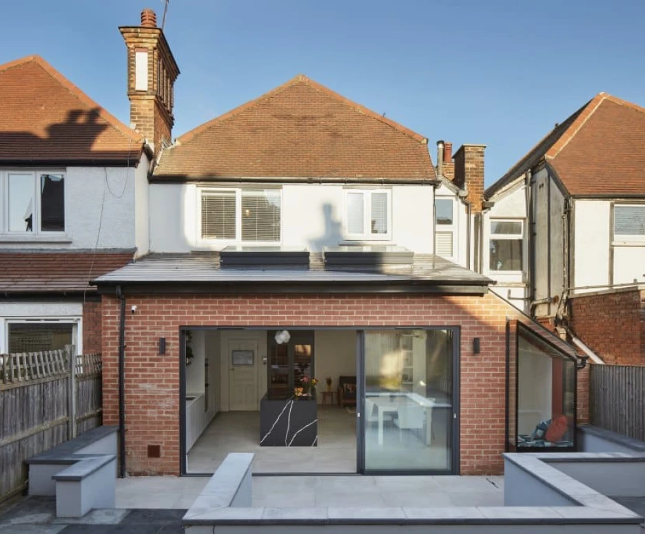 A modern two-story house with a brick exterior, featuring a large rear glass wall and a flat-roofed extension. The backyard includes a stone patio and decorative planters. Sunlight casts shadows from the chimney and adjacent houses.