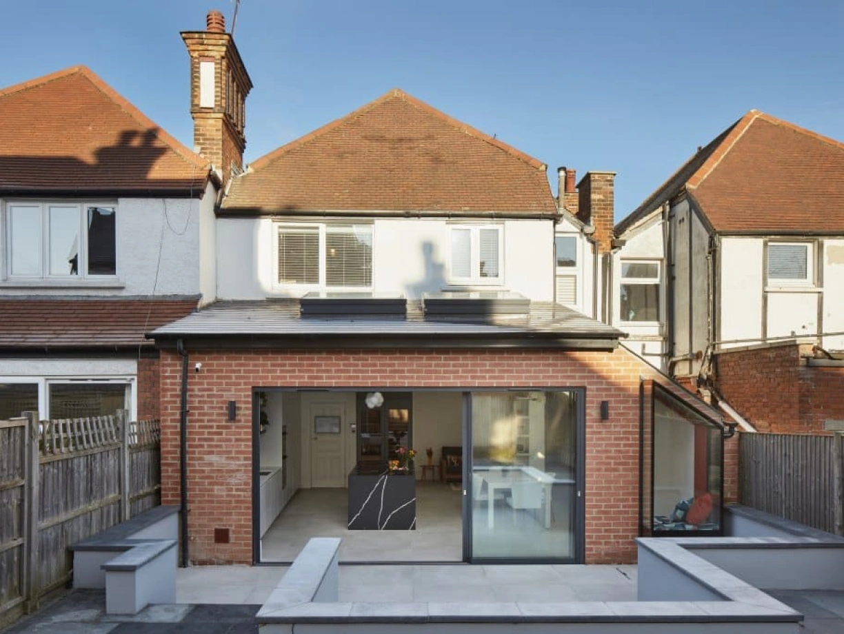 A modern two-story house with a brick exterior, featuring a large rear glass wall and a flat-roofed extension. The backyard includes a stone patio and decorative planters. Sunlight casts shadows from the chimney and adjacent houses.