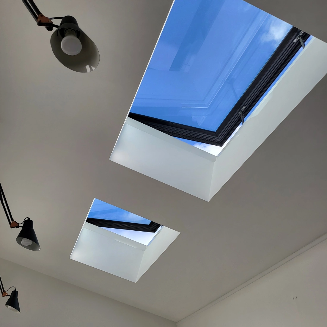 Two skylights in a modern ceiling allow natural light to enter, revealing a clear blue sky and few clouds. The ceiling is smooth and white, with sleek lighting fixtures along the sides.