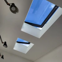 Two skylights in a modern room ceiling, framed by black borders, letting in natural light and revealing a clear blue sky. Adjustable pendant lights are mounted on the ceiling.
