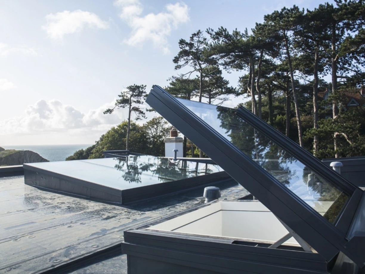 Skylights open on a flat roof, overlooking a scenic view of trees and the ocean under a partly cloudy sky.