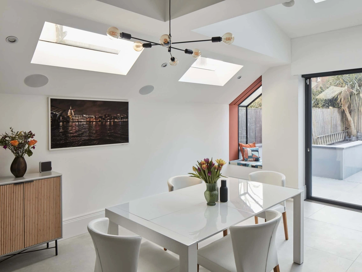 Bright modern dining area featuring a white table surrounded by four chairs. Skylights illuminate the space, complemented by a wall painting and a sideboard with a floral arrangement. A glass door leads to an outdoor patio.