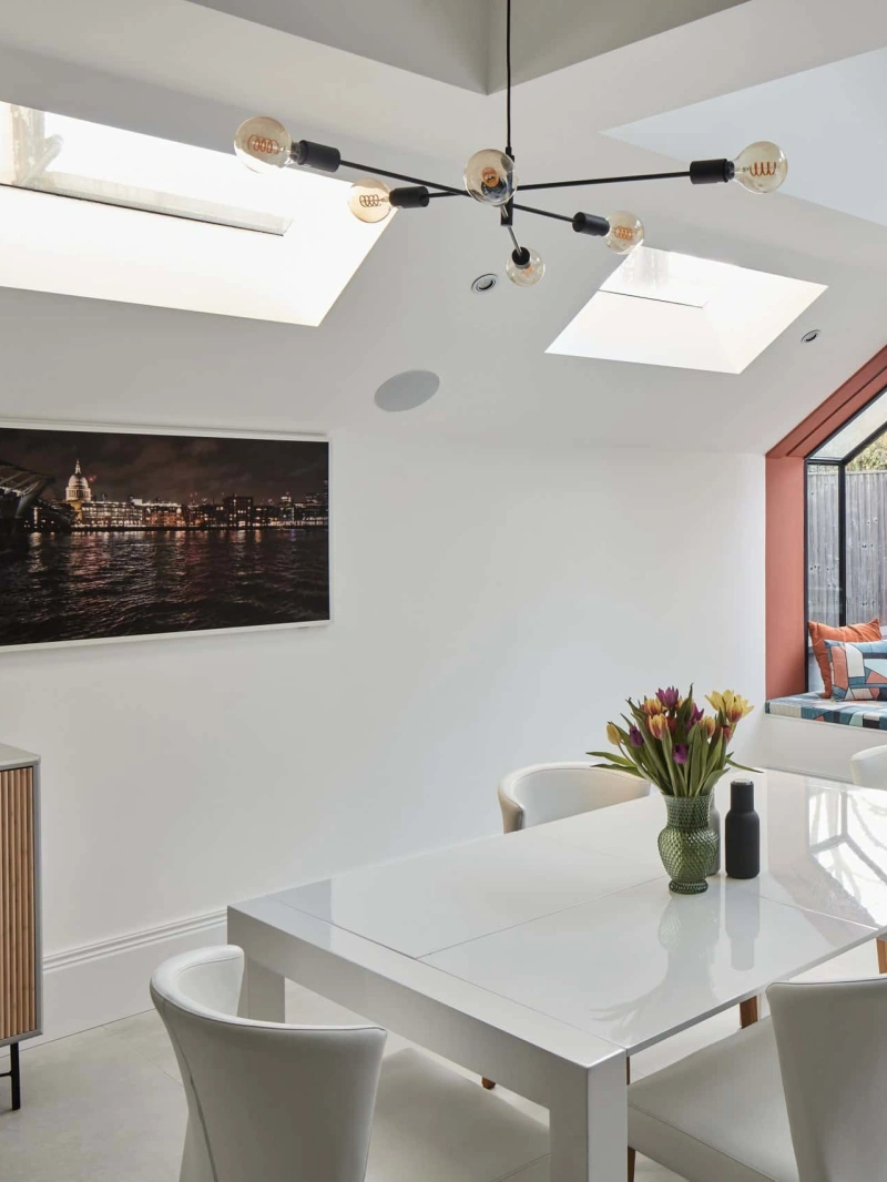 Bright modern dining area featuring a white table surrounded by four chairs. Skylights illuminate the space, complemented by a wall painting and a sideboard with a floral arrangement. A glass door leads to an outdoor patio.