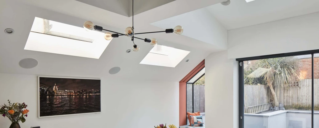 Bright modern dining area featuring a white table surrounded by four chairs. Skylights illuminate the space, complemented by a wall painting and a sideboard with a floral arrangement. A glass door leads to an outdoor patio.