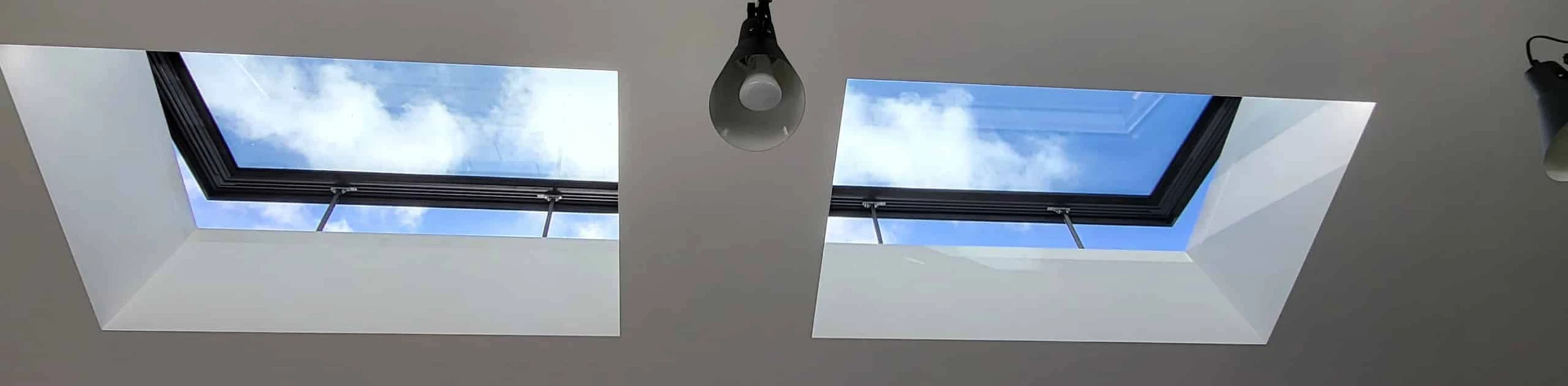 Two square skylights in a white ceiling, revealing a bright blue sky with clouds above. A light fixture hangs below one of the skylights.