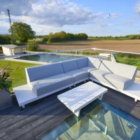 A modern outdoor seating area features a large gray sectional sofa and a white coffee table on a deck with glass panels. Lush greenery and fields extend in the background, under a partly cloudy sky.