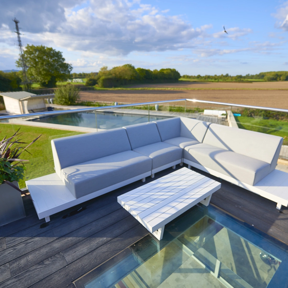 Un espace de détente extérieur avec un canapé gris en forme de L et une table basse blanche. En arrière-plan, un paysage de champs verdoyants sous un ciel nuageux.