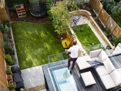 Vue aérienne d'un jardin aménagé avec du gazon et des plantes. Un homme se tient sur une terrasse, près d'un canapé moderne. On aperçoit un trampoline et un coin de jardin avec un petit banc et des plantations.