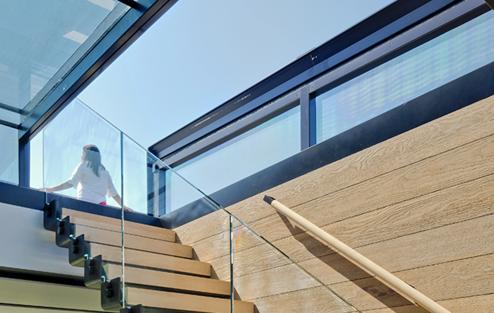 Escalier en bois clair avec rampe en verre, s'élevant vers un espace éclairé par de grandes fenêtres. Une personne monte les marches.