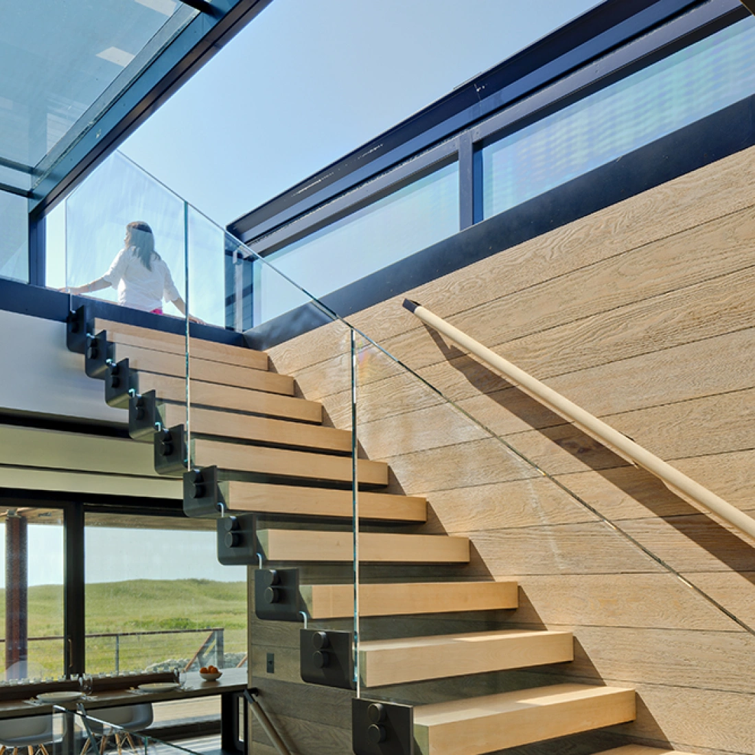 Escalier en bois clair avec rampe en verre, s'élevant vers un espace éclairé par de grandes fenêtres. Une personne monte les marches.