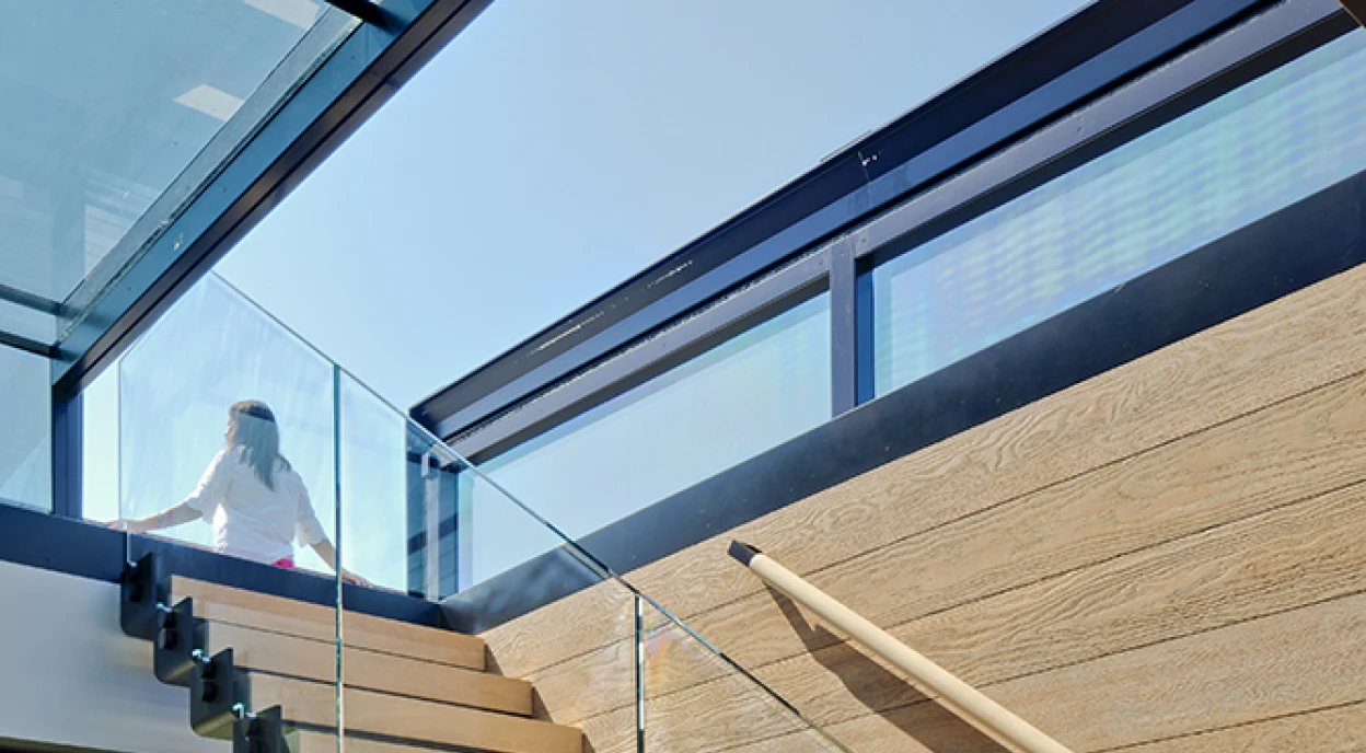 A modern staircase with wooden steps and glass railing, leading to a bright upper floor. Natural light floods the space, highlighting the contemporary design.