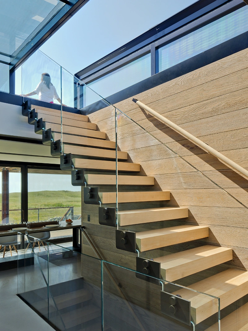 A modern staircase with wooden steps and a glass railing leads up to a bright, open space with large windows. A person is seen ascending the stairs, with sunlight streaming in from above.