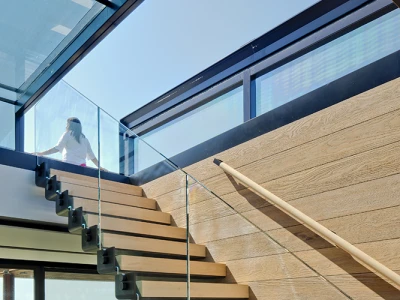 A modern staircase with wooden steps and glass railing, leading to a bright upper floor. Natural light floods the space, highlighting the contemporary design.