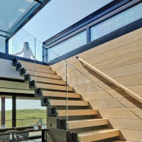 Escalera moderna con peldaños de madera y barandilla de vidrio. Al fondo, una persona se asoma por un ventanal que deja entrar luz natural. El ambiente es minimalista y contemporáneo.