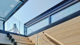 A modern staircase with wooden steps and glass railing, leading to a bright upper floor. Natural light floods the space, highlighting the contemporary design.