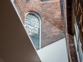 Ein Blick auf ein historisches Fenster aus buntverglastem Glas, eingebettet in rotem Mauerwerk, mit einem modernen architektonischen Element, das diagonal verläuft. Licht strömt durch das Fenster und beleuchtet den Raum.