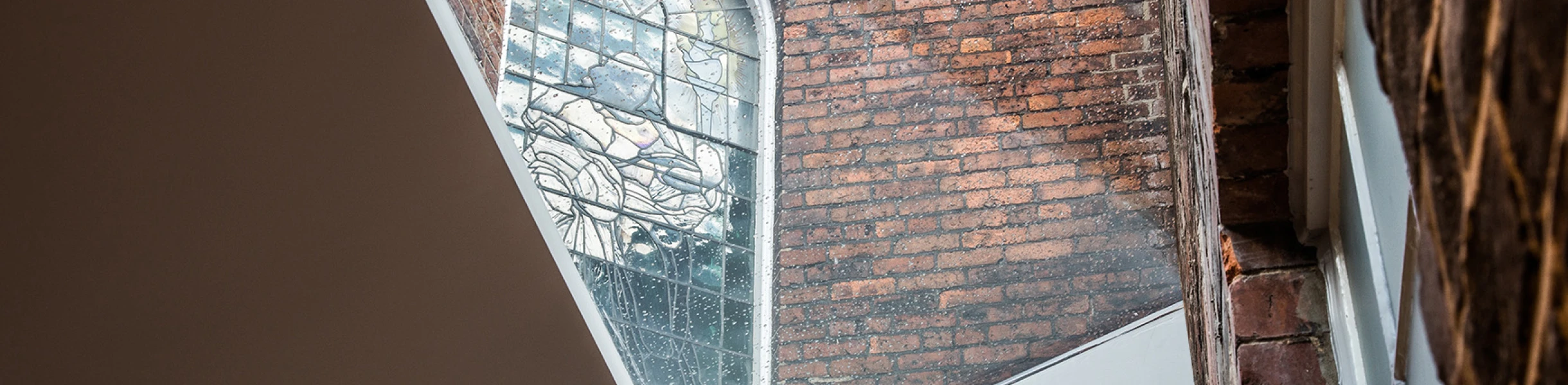 Ein Blick auf ein historisches Fenster aus buntverglastem Glas, eingebettet in rotem Mauerwerk, mit einem modernen architektonischen Element, das diagonal verläuft. Licht strömt durch das Fenster und beleuchtet den Raum.
