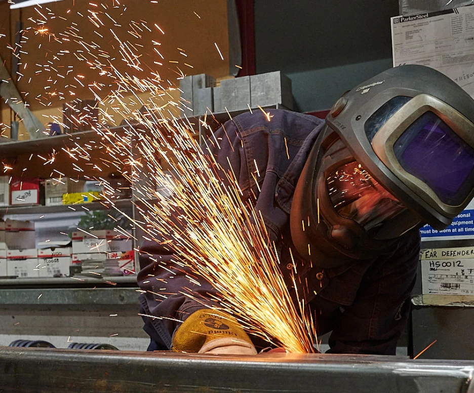 A welder in protective gear works on metal, creating bright sparks that illuminate the workshop. The background features shelves with various tools and materials.