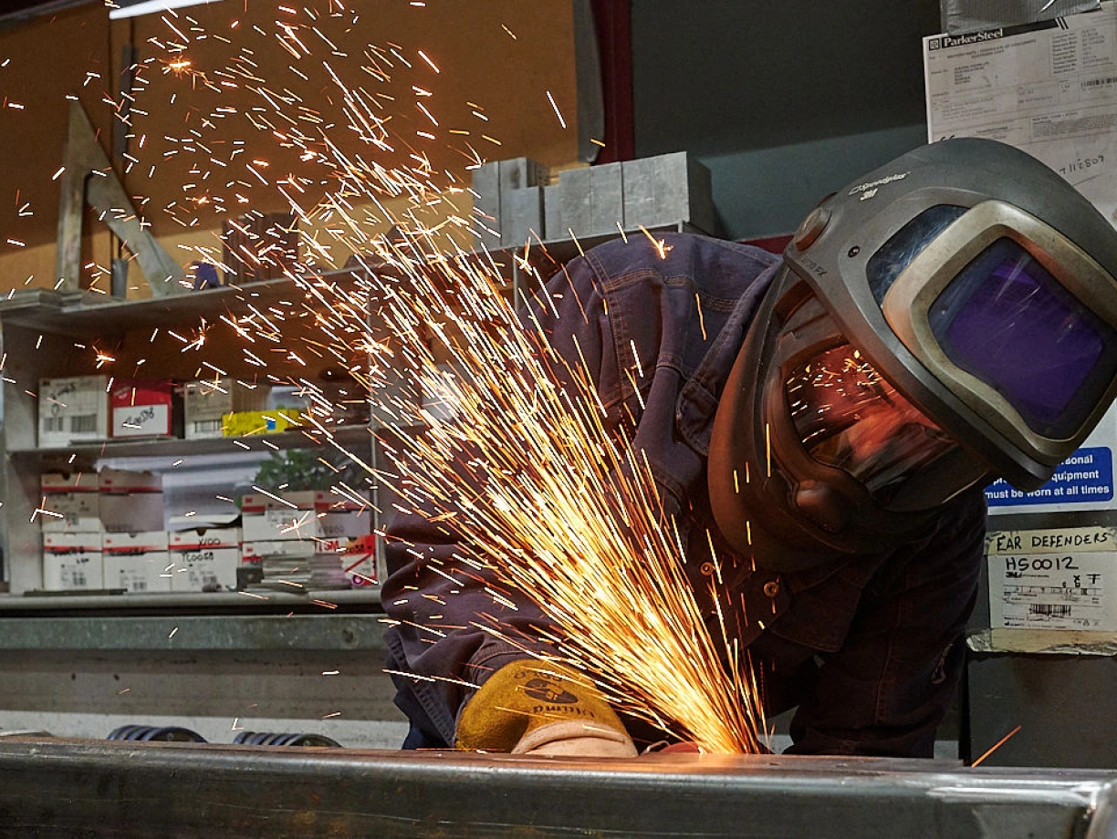 A welder in protective gear works on metal, creating bright sparks that illuminate the workshop. The background features shelves with various tools and materials.