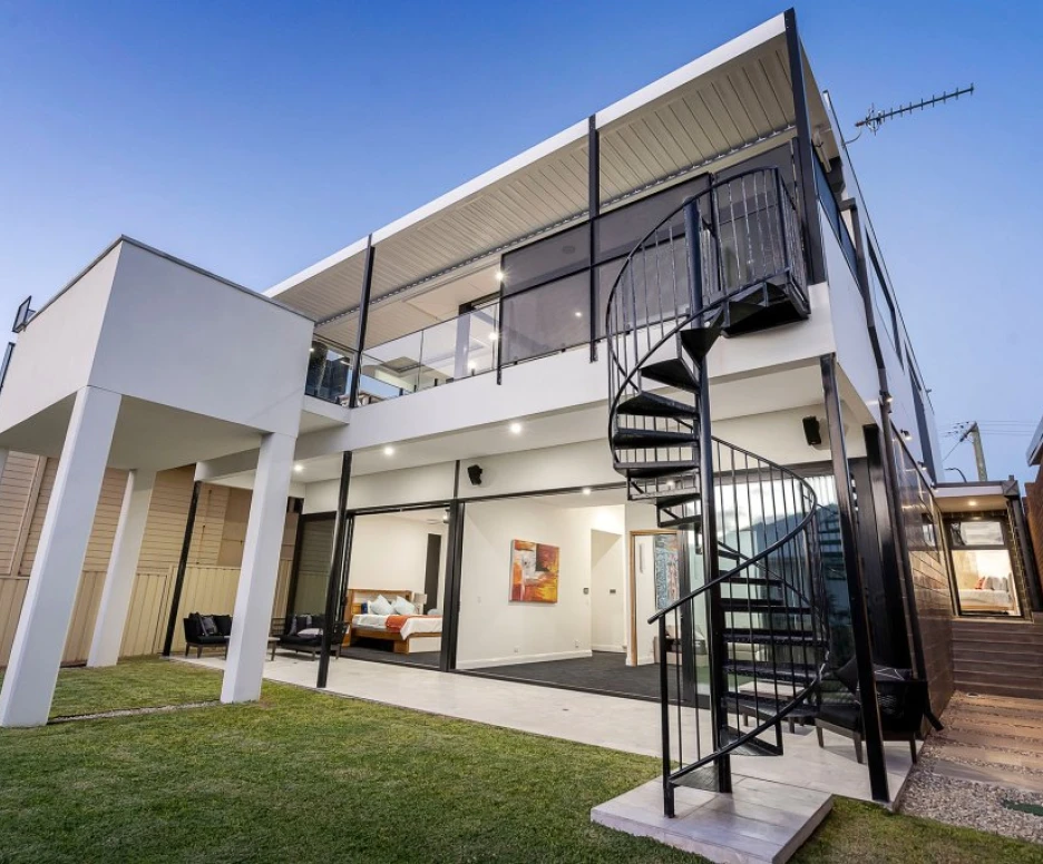 Maison moderne à plusieurs niveaux avec un escalier en colimaçon, un jardin bien entretenu et une verrière donnant sur l’intérieur.