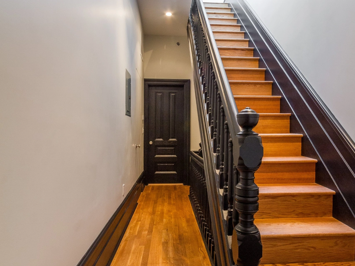 Narrow hallway leading to a staircase with wooden steps and a dark railing. Walls are light-colored, and there is a skylight above. A dark door is visible at the end of the hallway.