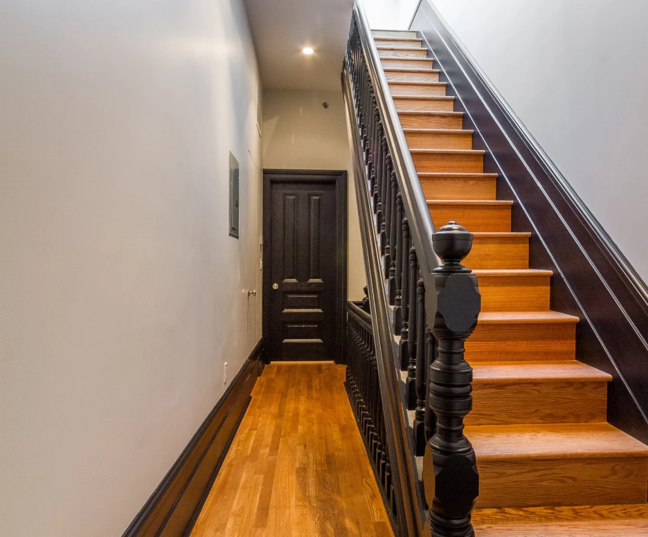 Narrow hallway leading to a staircase with wooden steps and a dark railing. Walls are light-colored, and there is a skylight above. A dark door is visible at the end of the hallway.