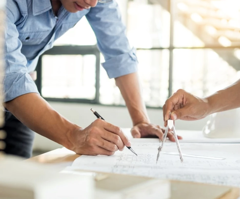 Deux personnes travaillent sur des plans architecturaux. L'une utilise un compas pour tracer des lignes, tandis que l'autre prend des notes avec un stylo. Un casque de sécurité est visible sur la table. La lumière naturelle entre par les fenêtres en arrière-plan.