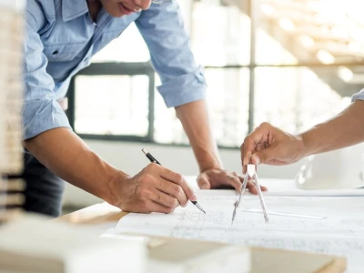 Deux personnes travaillent sur des plans architecturaux. L'une utilise un compas pour tracer des lignes, tandis que l'autre prend des notes avec un stylo. Un casque de sécurité est visible sur la table. La lumière naturelle entre par les fenêtres en arrière-plan.