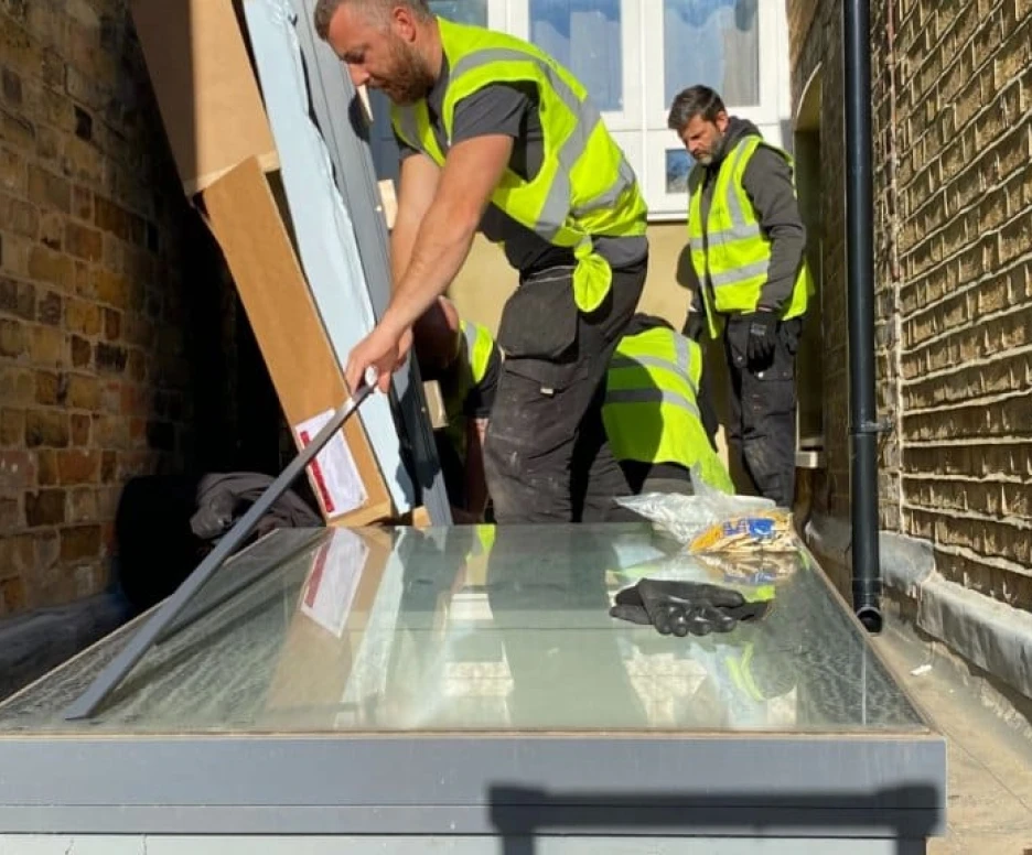 Workers in safety vests install a large glass panel on a paved area beside a brick wall. Tools and materials are scattered around the workspace.