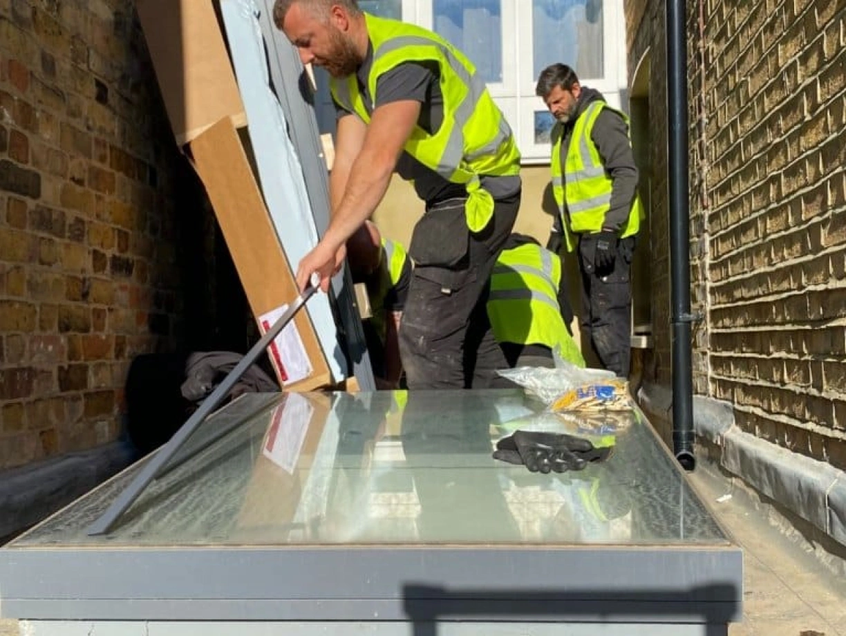 Workers in safety vests install a large glass panel on a paved area beside a brick wall. Tools and materials are scattered around the workspace.