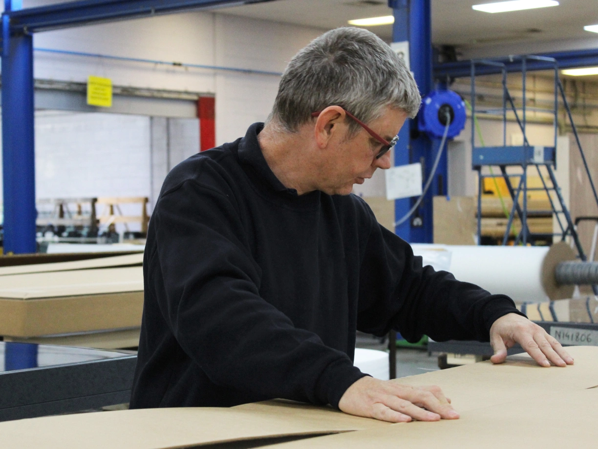 Un homme aux cheveux gris, portant des lunettes, travaille sur une surface en carton dans un environnement d’atelier. Il se penche en avant, concentré sur sa tâche.