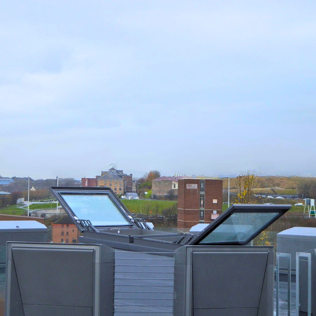 A rooftop view featuring open skylights, with a background of buildings and greenery under a cloudy sky.