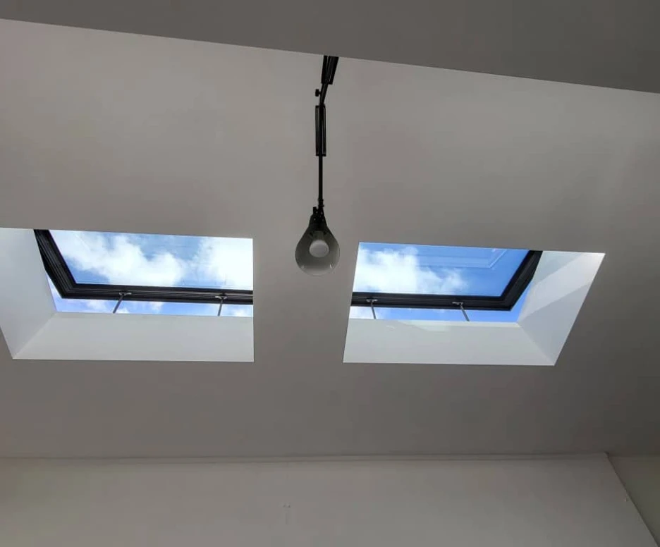 Two skylights set into a white ceiling, showcasing a blue sky with fluffy clouds. A pendant light hangs between the skylights.