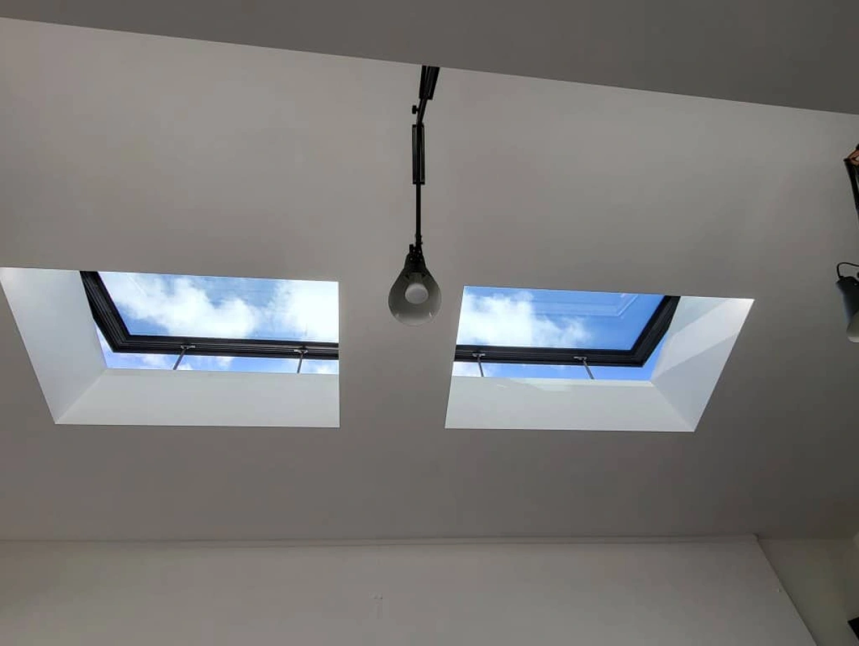 Two skylights set into a white ceiling, showcasing a blue sky with fluffy clouds. A pendant light hangs between the skylights.