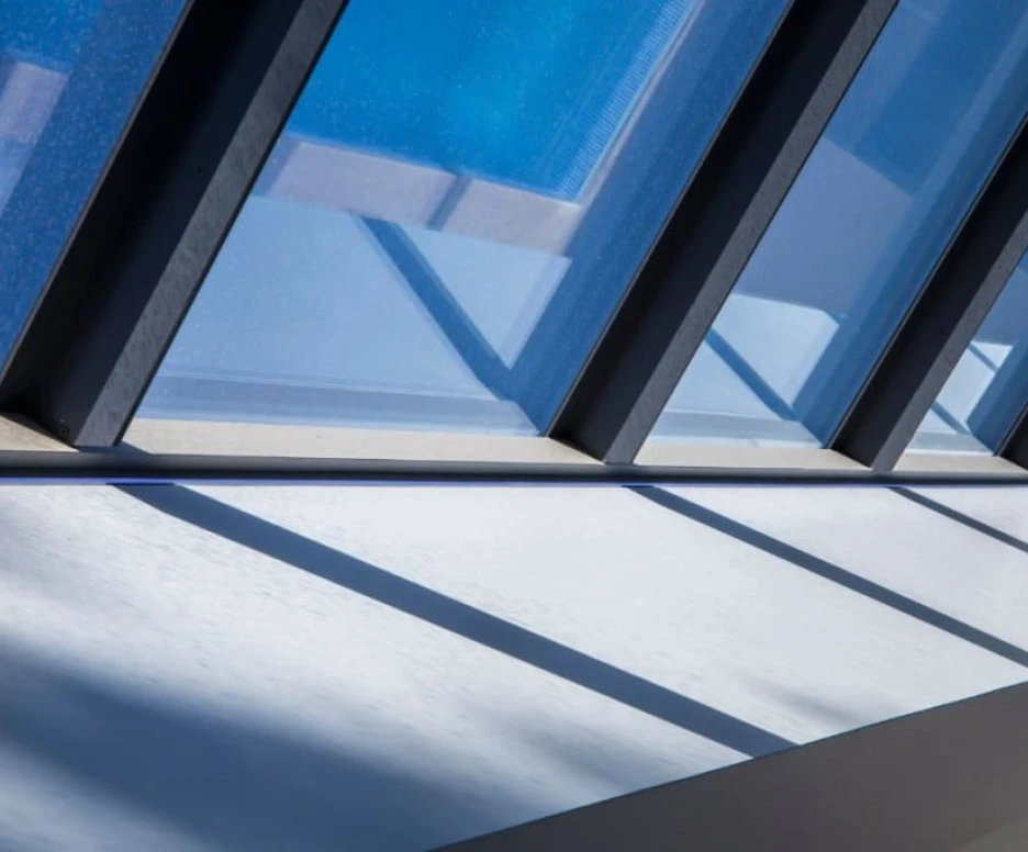 Sunlight filters through a series of angled windows, casting geometric shadows on a white surface below. The blue sky is reflected in the glass, creating a tranquil and modern aesthetic.