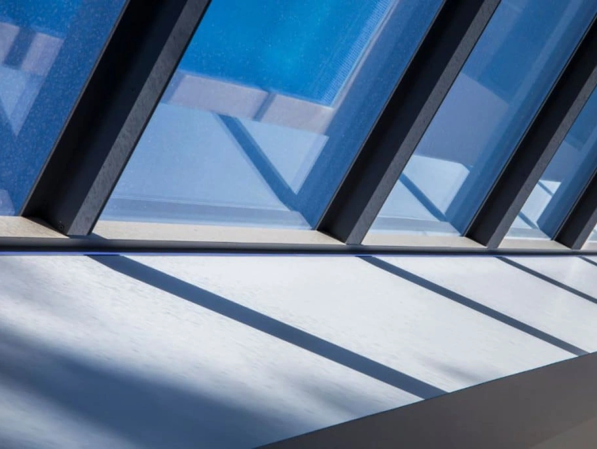 Sunlight streams through large, angled glass panels, casting geometric shadows on a white surface below. The clear blue sky is visible in the background.