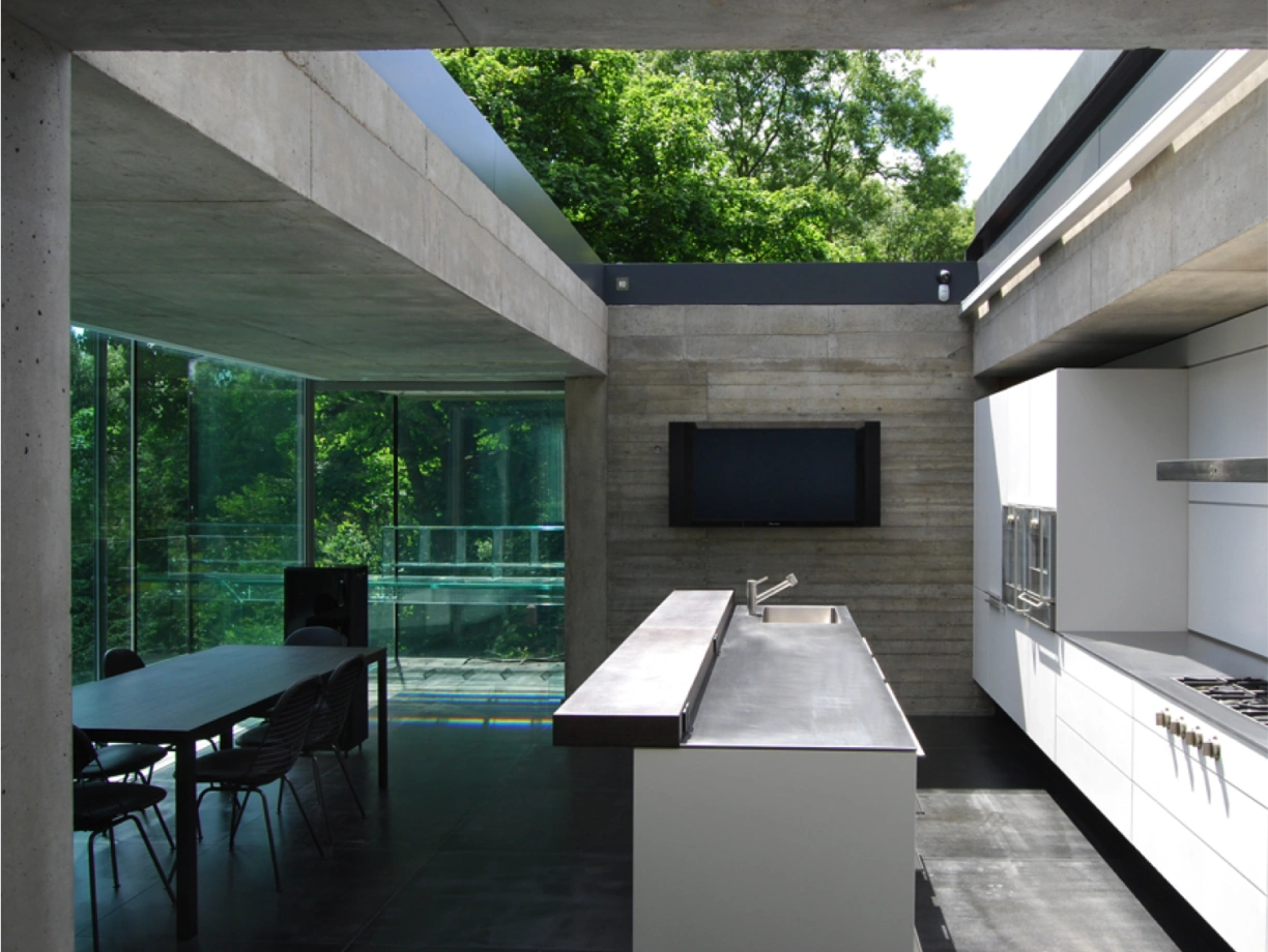 Cocina moderna con paredes de hormigón y grandes ventanales que dan a un entorno natural. Hay una mesa de comedor de madera y sillas negras, y un espacio brillante y minimalista con electrodomésticos de acero inoxidable.