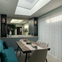 Une salle à manger moderne avec une table au centre, entourée de chaises. Un plafond en verre laisse entrer la lumière, tandis qu'un canapé turquoise est sur le côté. Des vases et des décorations ajoutent une touche élégante.