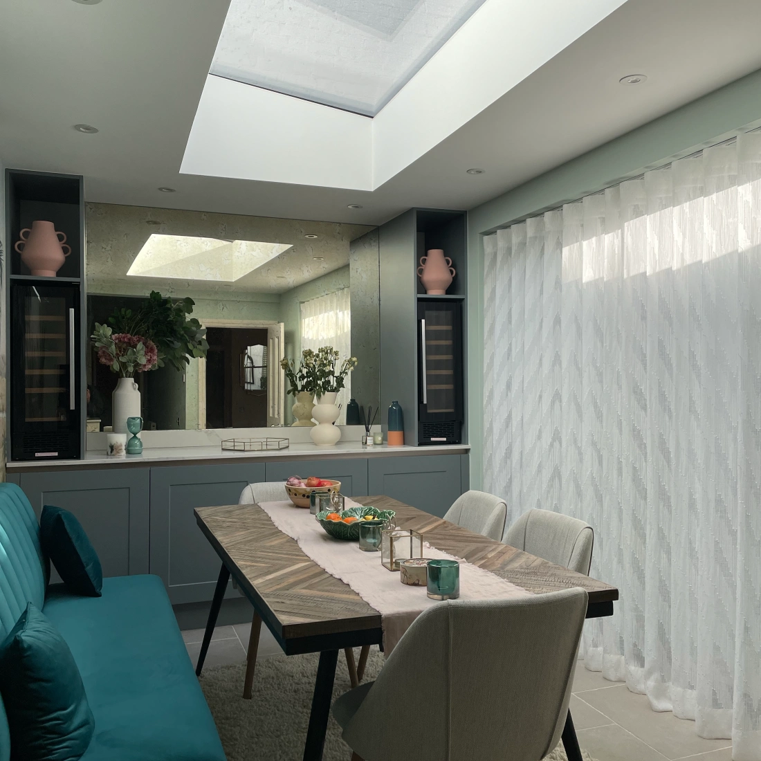 A modern dining area featuring a light wood table set with decorative items and a pastel runner. Surrounding are six light-colored chairs. A skylight above fills the space with natural light, while sheer curtains soften the ambiance. Green plants and decorative vases add a touch of color to the stylish decor.