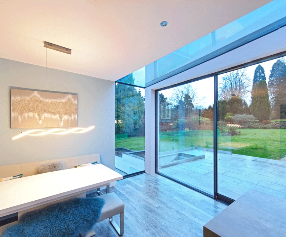 A modern dining area featuring a sleek white table with comfortable seating, large glass windows offering a view of a landscaped garden, and a contemporary light fixture hanging above the table. The space has a combination of light tones and textures, creating a bright and inviting atmosphere.