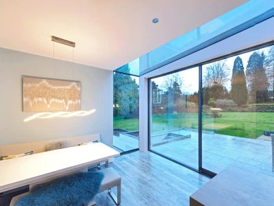 Un intérieur moderne d’une salle à manger avec une grande fenêtre donnant sur un jardin verdoyant. Une table blanche élégante est entourée de sièges confortables. Des lumières suspendues ajoutent une ambiance chaleureuse.