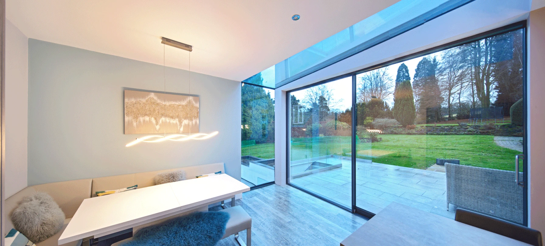 Moderner Essbereich mit einem großen Fenster, das auf einen Garten mit Bäumen und Rasenblick gibt. Ein eleganter Tisch mit vier Stühlen ist im Vordergrund, und eine schlichte Lampe hängt über dem Tisch. Der Raum hat entspannte Farben und natürliches Licht.